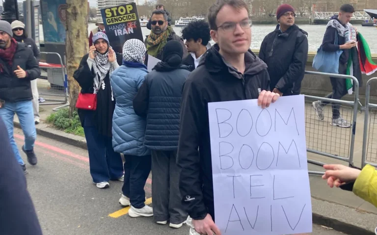 Pro-Iran protesters holding a sign that reads 'boom boom Tel Aviv' at a London rally, with a crowd of people in the background and a mix of colours from the protesters' signs and banners