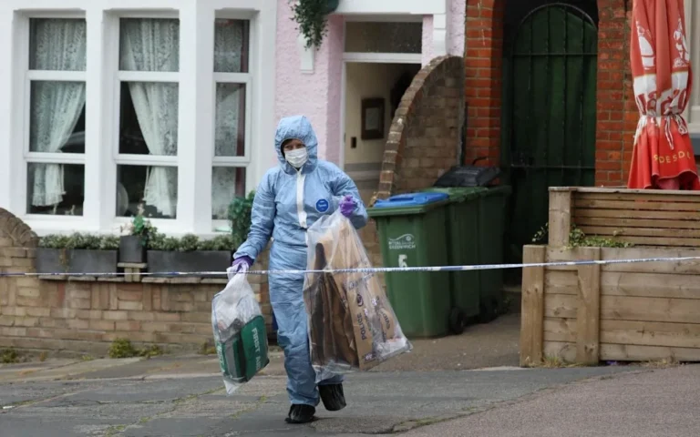 A forensic officer examining the scene outside the elderly woman's home, where she was fatally stabbed, with police tape and officers in the background, highlighting the seriousness of the incident and the need for community safety