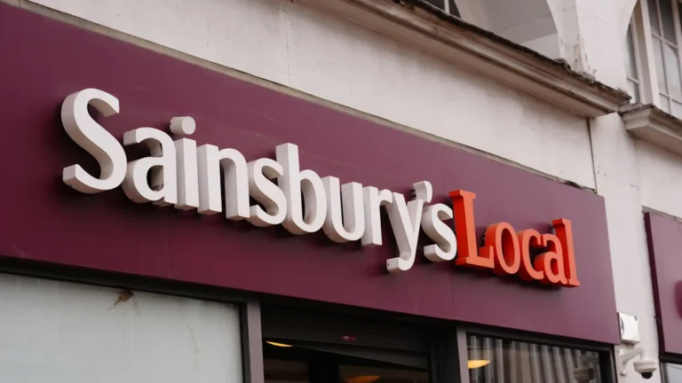 A Sainsbury's store front with a sign reading 'open for business' as people walk in and out, symbolising the company's resilience amid the Iran war, with a focus on consumer behaviour and economic impact