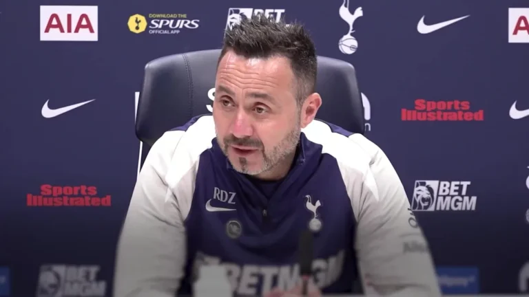 Roberto De Zerbi, the manager of Tottenham Hotspur, is seen giving a team talk to his players, emphasizing the importance of victory and a winning mentality, with the team's colours and logo prominently displayed, symbolizing their collective effort and behaviour on the pitch