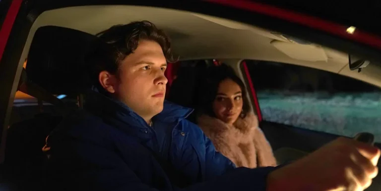 Hollyoaks' Charlie Dean stands amidst the wreckage of a car crash, looking shocked and accusatory, with a mix of colours and debris surrounding the scene, as the primary keyword 'car crash' is evident in the aftermath