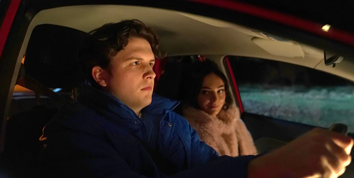 Hollyoaks' Charlie Dean stands amidst the wreckage of a car crash, looking shocked and accusatory, with a mix of colours and debris surrounding the scene, as the primary keyword 'car crash' is evident in the aftermath