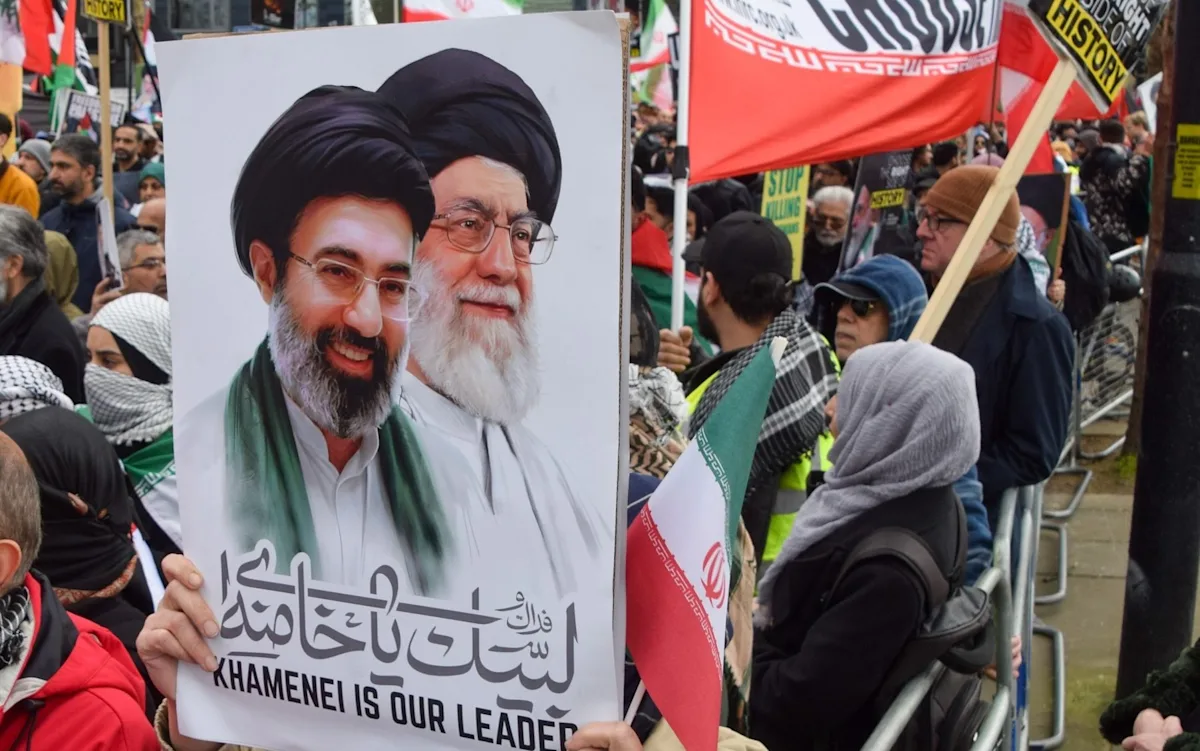 A group of people gathered around a microphone, with a banner in the background reading 'Free Speech', highlighting the complex issue of Iranian propagandists being given a platform by British charities, and the need to balance free speech with the prevention of harm, in the context of social justice and charitable responsibility