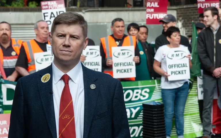 A crowded train station with commuters waiting for trains, with a sign in the background reading 'train strikes' and a union flag, highlighting the RMT union's threat of maximum disruption to the UK's rail network