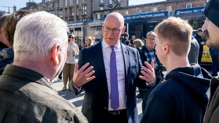 A rural village in the UK with a group of people standing outside a local shop, highlighting the issue of rural costs and the need for government action to support rural communities