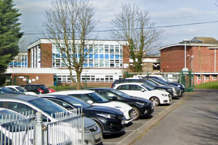 A school teacher standing in a classroom with a concerned expression, with a blurred background of students in the foreground, highlighting the importance of school safety and student wellbeing in the wake of an attempted murder incident