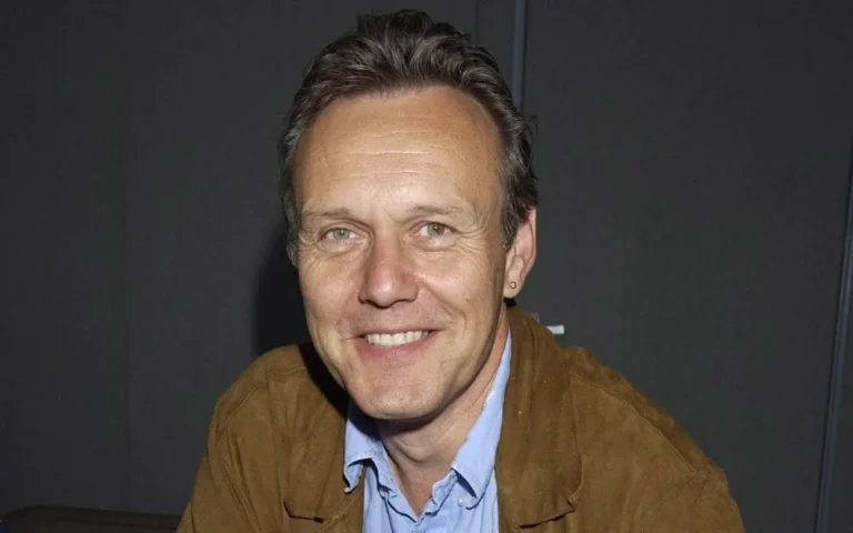 Anthony Head, the Gold Blend man, sitting on a couch with a cup of coffee, looking charming and charismatic, with a warm and colourful background, showcasing his behaviour and personality