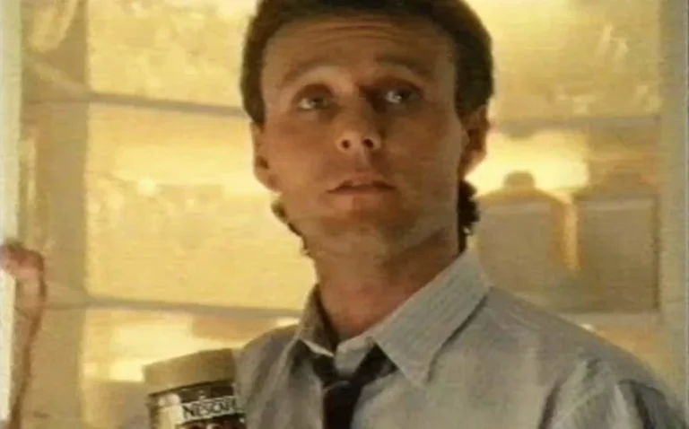 Anthony Head as the Gold Blend man, sitting on a couch with a cup of coffee, looking charming and sophisticated, with a warm colour palette and a relaxed atmosphere, showcasing his iconic role in the 1980s coffee adverts