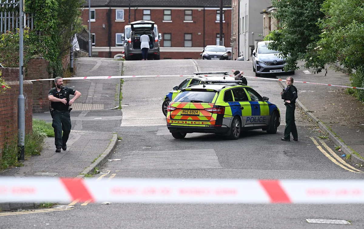 A photo of a police car in Belfast, with a Sudanese migrant in the background, highlighting the issue of community safety and migrant integration, with a focus on the alleged attempted beheading incident