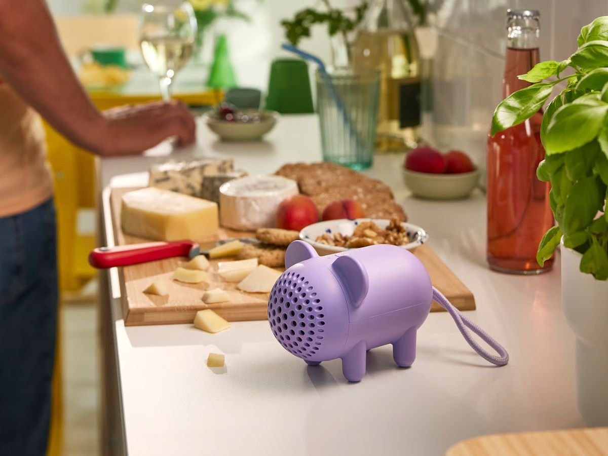The IKEA Grejsimojs Portable Bluetooth speaker, featuring a purple mouse-shaped design with ears and a tail, on a wooden table surrounded by plants and books, showcasing its compact size and colourful appearance, perfect for indoor or outdoor use, with a primary focus on portable Bluetooth speakers