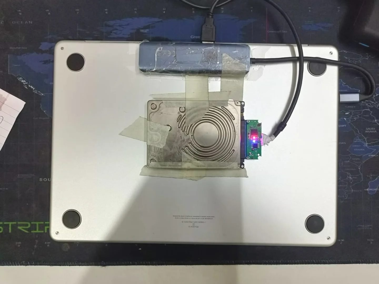 A photo of the M2 MacBook Air with a USB-C dongle and SATA drive attached using low-quality tape, showcasing the creative mod to combat rising SSD prices, with the primary keyword 'SSD prices' featured prominently