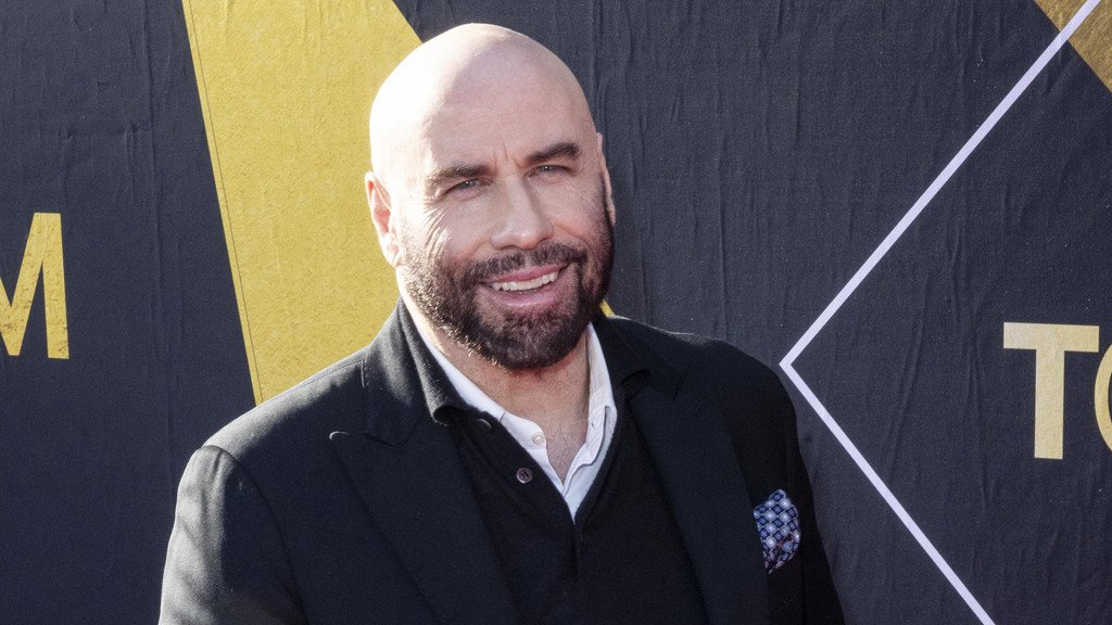 John Travolta standing in front of a private jet, smiling and holding his new licence, with a clear blue sky in the background, highlighting his love for aviation and flying