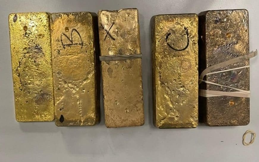 A pile of gold bars with a police officer in the background, symbolising the seizure of £1.8m worth of gold at an airport, highlighting the importance of airport security and the fight against money laundering
