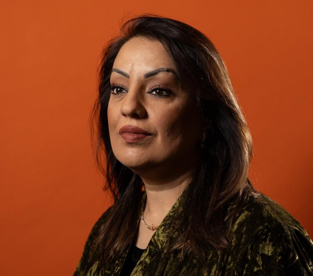 Labour MP Naz Shah speaking at a community event, with a determined look on her face, highlighting her journey towards healing and growth after a murder arrest, with a mix of colour and emotion in the background