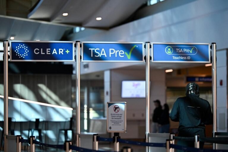 A UK airport security checkpoint with a sign indicating the suspension of TSA PreCheck and Global Entry programmes, with travellers waiting in line, highlighting the impact on airport security and travel