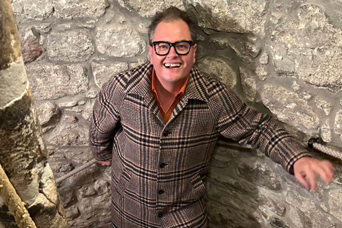 Alan Carr standing in front of his newly purchased Scottish castle, surrounded by rolling hills and colourful scenery, with a hint of his new tartan, showcasing his excitement and joy at becoming a part of Scottish culture and heritage