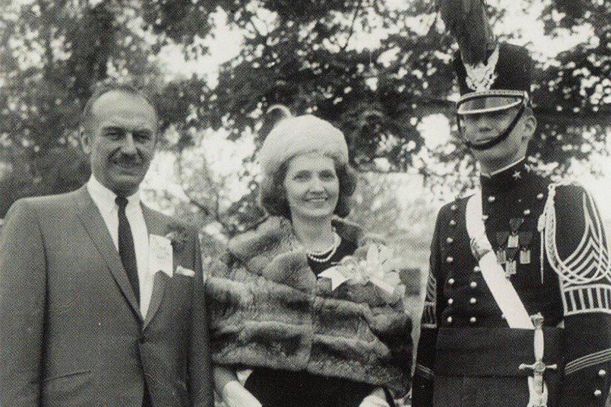 Donald Trump sitting in the Oval Office, surrounded by American flags and military personnel, with a picture of his parents on his desk, symbolising his admiration for the American military and its dominance, a topic of significant interest and discussion