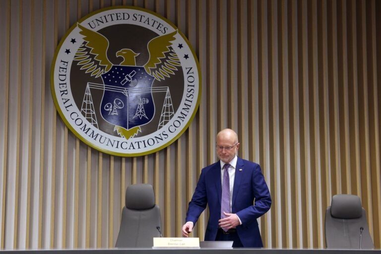 A photo of a person holding a sign that reads 'Free Speech' with a blurred background of people protesting, highlighting the importance of freedom of speech and the current threats to it, with a focus on the FCC chair's recent actions