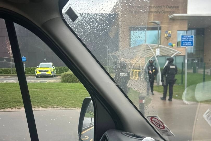 Armed police officers outside a school building, with students and parents looking on in concern, highlighting the need for increased security measures to ensure student safety and prevent school closures