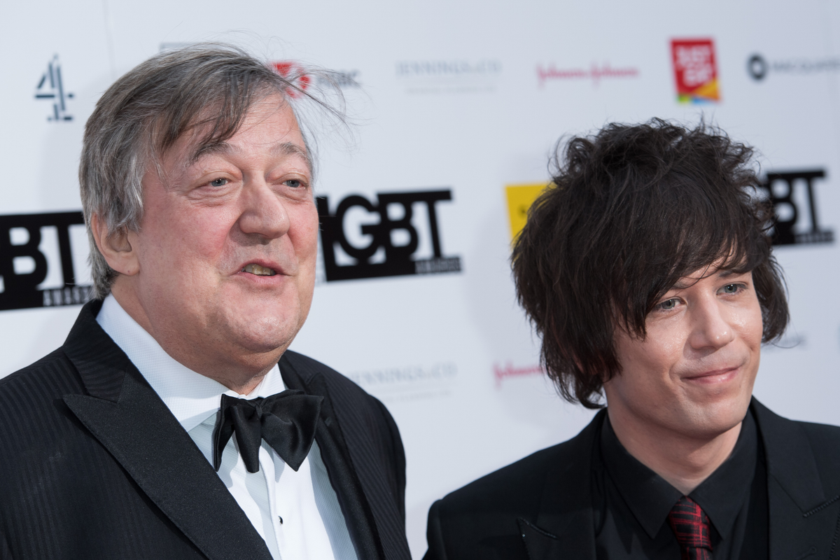 Stephen Fry and Elliott Spencer, a couple with a significant age gap, smiling and embracing each other, surrounded by a warm and loving atmosphere, with a beautiful colour palette in the background, highlighting the beauty of their love and commitment to each other