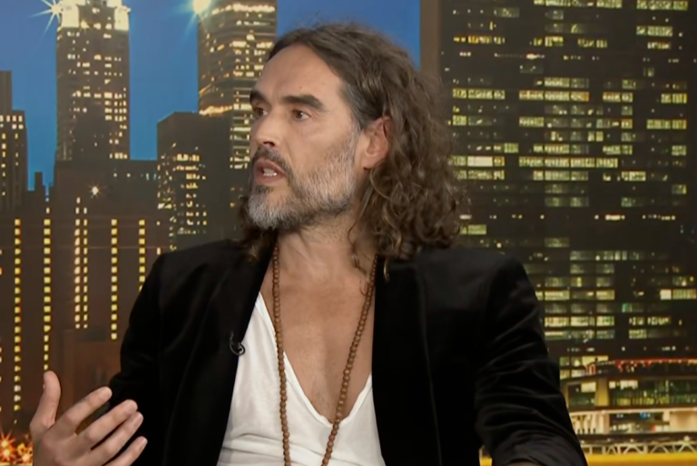 Russell Brand looking embarrassed during his Piers Morgan interview, struggling to find a Bible passage, with a mixture of shock and amusement on his face, showcasing his behaviour during the painful conversation