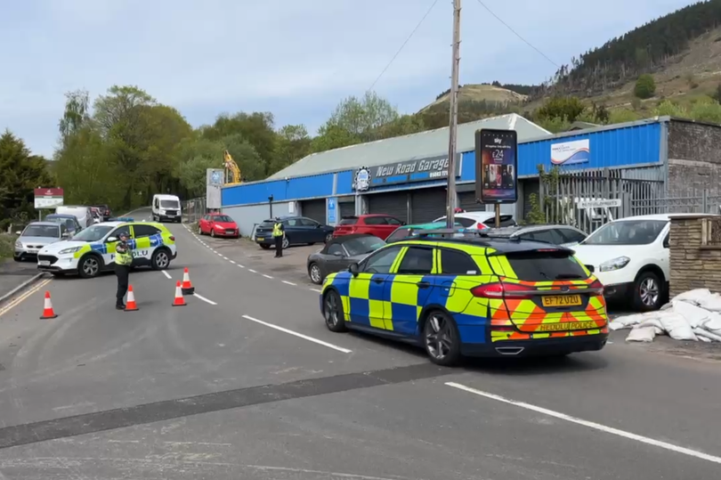 Aerial view of the Welsh mountain with a large crowd of people gathered for the illegal rave, with police cars and ambulances in attendance, highlighting the need for public safety and environmental protection