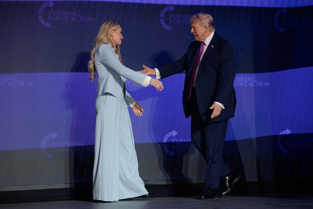 Erika Kirk standing on stage with Donald Trump at the Turning Point rally, with a crowd of people in the background, as she introduces him amidst a flurry of security concerns and controversy surrounding her recent decision to skip a Vance event due to serious threats, with her behaviour and priorities under scrutiny