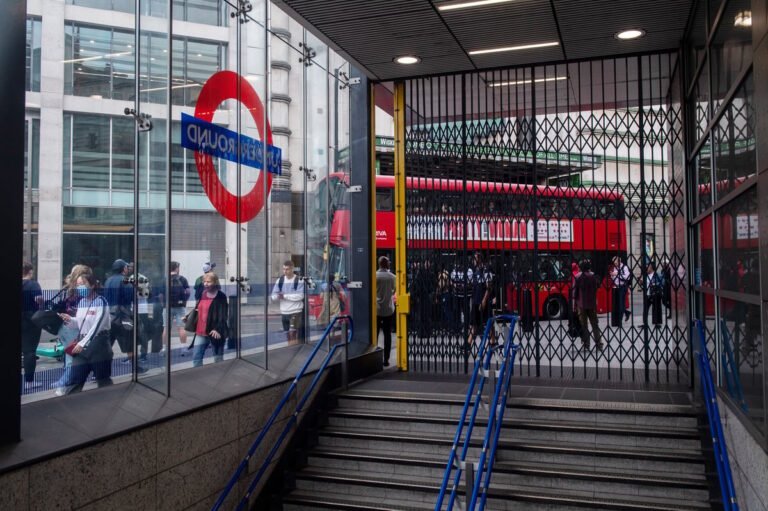 London tube strike chaos: commuters wait at a busy station as RMT union members protest outside, holding signs that read 'fair pay for fair work' and 'support our cause', with a colourful backdrop of London's bustling streets