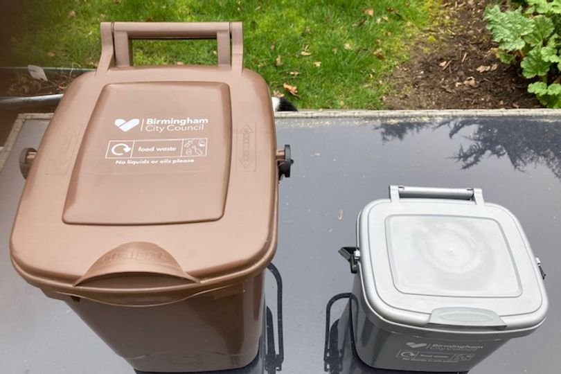 A binman standing next to a food waste caddy with a red cross through it, highlighting the importance of proper waste disposal and the consequences of contamination, with a focus on food waste management