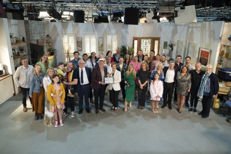 Neighbours cast members standing outside the fictional Ramsay Street, with a mix of happy and sad expressions, as the show finds a new home in the UK after Amazon Freevee cancellation, with a red door and colourful flowers in the background, symbolising the show's iconic theme and setting