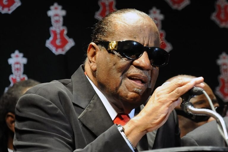Clarence Carter performing on stage, singing his hit song Strokin, with a soulful voice and energetic behaviour, surrounded by colour and music