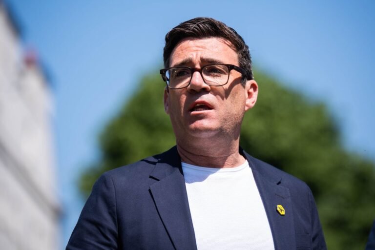 Andy Burnham speaking at a political rally, with a crowd of supporters in the background, waving flags and banners with the Labour party colour scheme, as he campaigns to beat Reform in Makerfield and take on Starmer