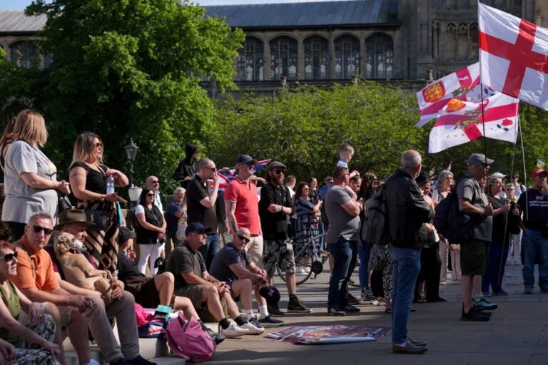 A group of protesters holding signs and banners in Norfolk, with a police presence in the background, following the arrest of seven refugees charged with child sexual offences, highlighting the need for stricter vetting and integration policies