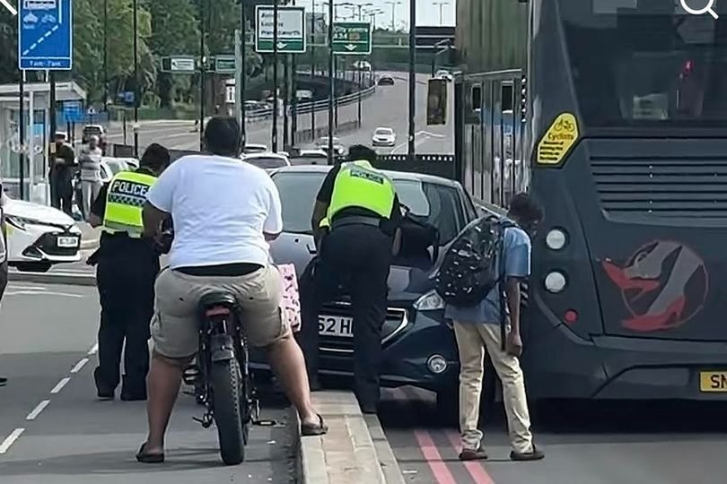 A car trapped beside a bus on a busy Birmingham route, with people gathered around, trying to make sense of the situation, the primary keyword being 'Birmingham bus route'