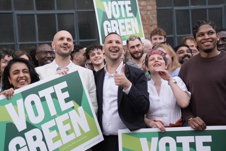 The Green Party's leader speaking at a London town hall meeting, surrounded by supporters and campaigners, with a focus on the Gaza ban and environmental issues