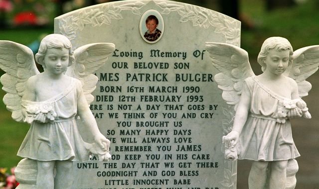 A photo of a grave with flowers and a damaged headstone, with a blur of people in the background, the primary keyword phrase is James Bulger grave vandalism, the image depicts a sombre mood and a sense of respect for the deceased, the colour of the grave is a dull grey, the overall appearance is one of sadness and loss